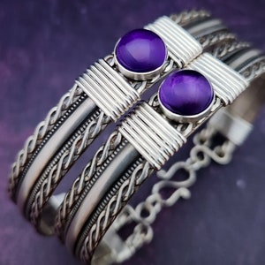 May include: Two silver-toned bracelets with intricate braided and wire-wrapped designs. Each bracelet features a large, round, vibrant purple gemstone. The bracelets are displayed against a blurred purple background.