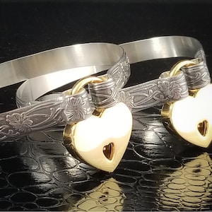 Puede incluir: Dos pulseras de plata con acabado oxidado negro y dijes de corazón con cerradura de oro. Las pulseras están grabadas con un diseño floral.