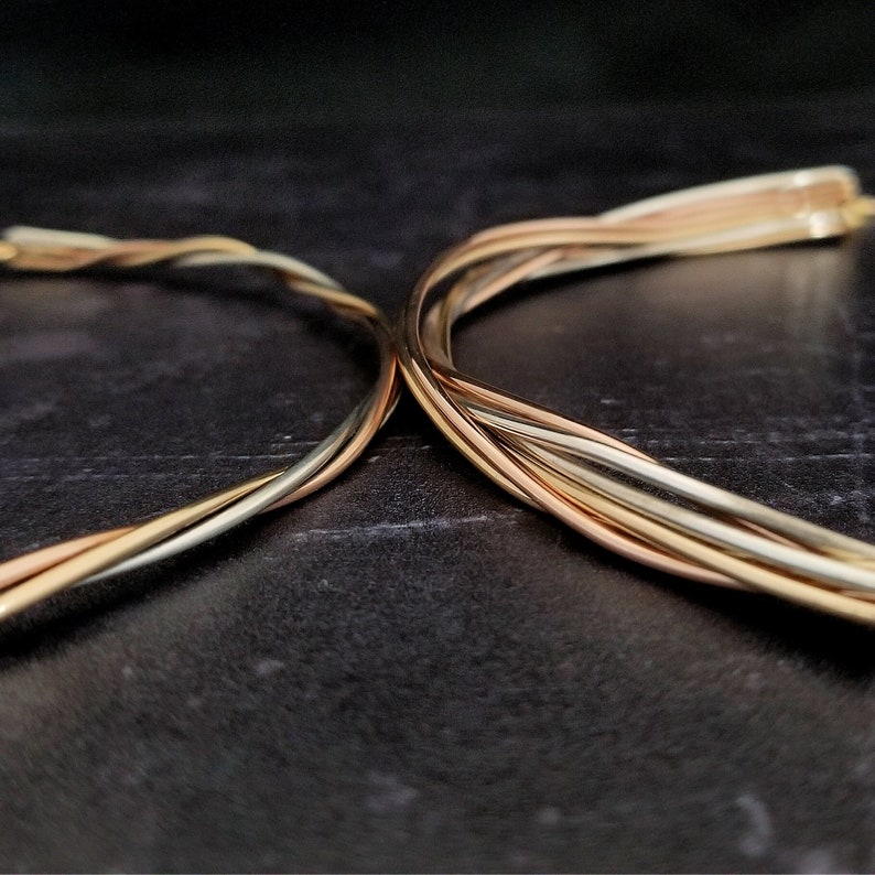 May include: Three strands of wire, one gold, one silver, and one rose gold, are twisted together to form a decorative piece. The wires are arranged in a criss-cross pattern, creating a visually appealing design.