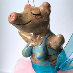 May include: A ceramic pig figurine dressed as a ballerina, wearing a pink tutu and a blue and yellow bra. The pig has a pink snout and is standing on its hind legs.