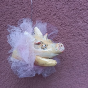 May include: A ceramic pig ornament wearing a pink tulle tutu. The pig has a white face with painted eyes and a pink snout.