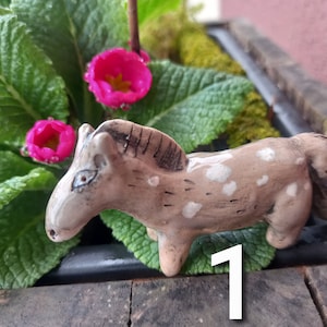 Puede incluir: Una peque&ntilde;a figurita de caballo de cer&aacute;mica pintada a mano con manchas marrones y blancas. El caballo est&aacute; de pie sobre una superficie marr&oacute;n con hojas verdes y flores rosas en el fondo.