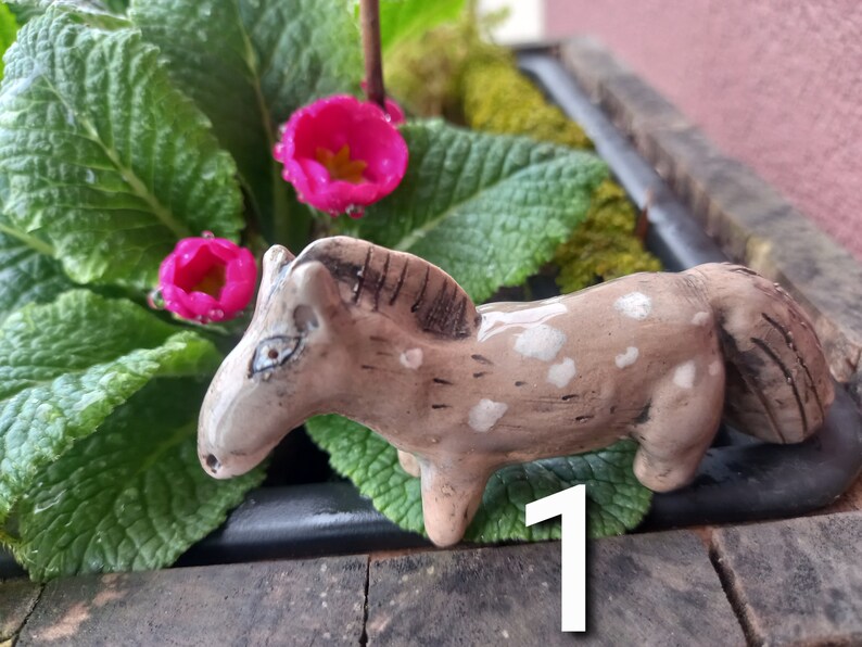 Puede incluir: Una peque&ntilde;a figurita de caballo de cer&aacute;mica pintada a mano con manchas marrones y blancas. El caballo est&aacute; de pie sobre una superficie marr&oacute;n con hojas verdes y flores rosas en el fondo.
