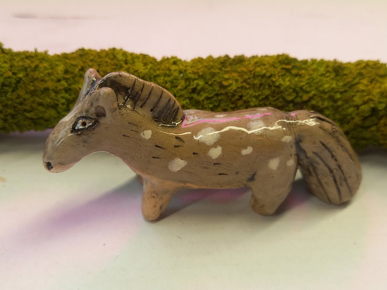 Puede incluir: Una peque&ntilde;a figurita de caballo de cer&aacute;mica con pintura moteada marr&oacute;n y blanca. El caballo tiene una cola y una crin largas.