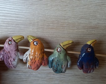 May include: Four ceramic bird figurines with yellow beaks and painted details. The birds are pink, orange, green, and blue. They are arranged in a row on a wooden surface.