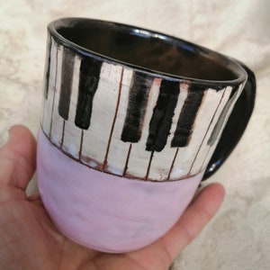 May include: A ceramic mug with a black and white piano key design. The mug is painted with a pink glaze on the bottom half.