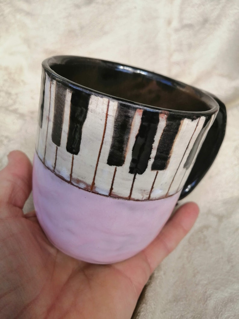 May include: A ceramic mug with a black and white piano key design. The mug is painted with a pink glaze on the bottom half.