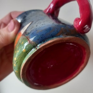 May include: A handmade ceramic mug with a red handle and a colourful, glazed exterior. The mug has a green, blue, and orange design with a red interior.