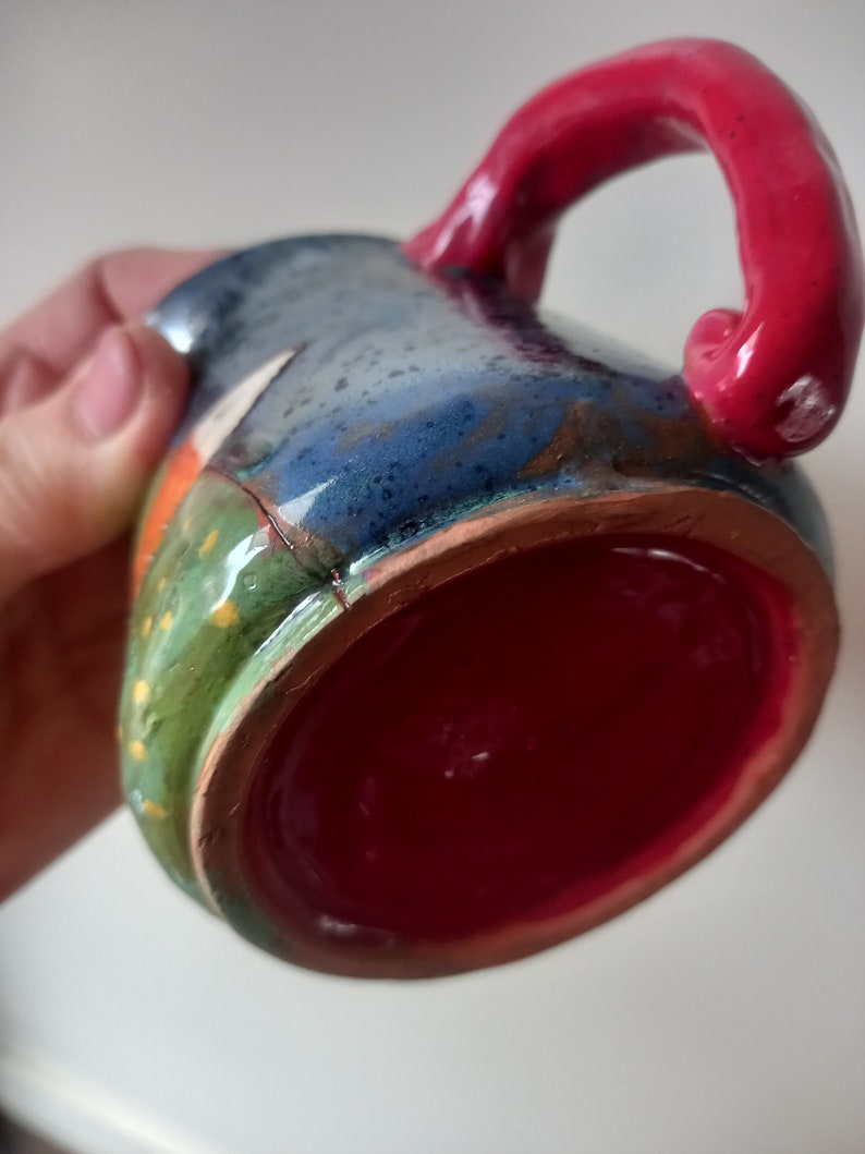 May include: A handmade ceramic mug with a red handle and a colourful, glazed exterior. The mug has a green, blue, and orange design with a red interior.