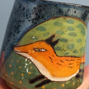 May include: A ceramic mug featuring a hand-painted design of a fox leaping over a green hill with yellow and green spots. The mug has a blue glaze with a speckled finish.