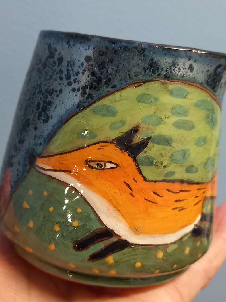 May include: A ceramic mug featuring a hand-painted design of a fox leaping over a green hill with yellow and green spots. The mug has a blue glaze with a speckled finish.