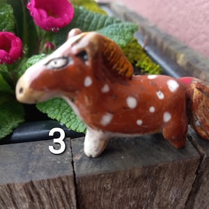 Puede incluir: Una figurita de caballo de cer&aacute;mica marr&oacute;n y blanca con manchas blancas. El caballo tiene una crin y una cola marrones. La figurita est&aacute; sentada sobre una superficie de madera. El n&uacute;mero "3" es visible en la imagen.