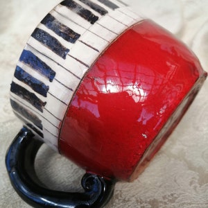 May include: A red ceramic mug with a black handle. The mug is decorated with a black and white piano keyboard design.