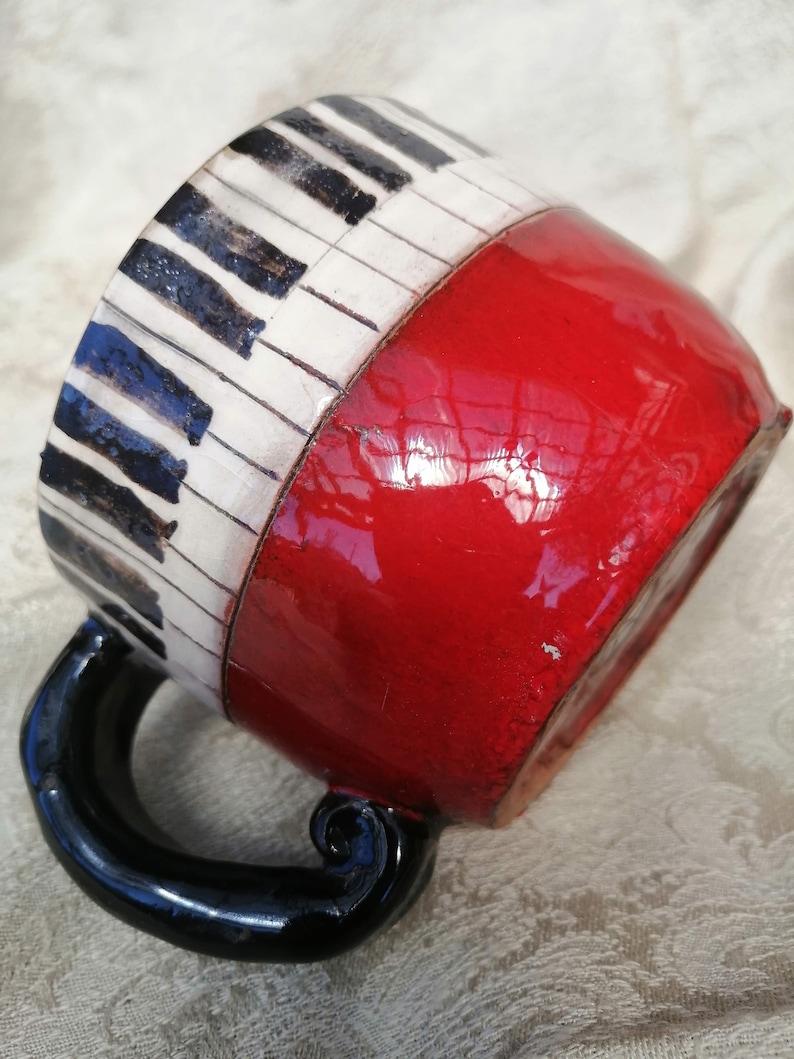 May include: A red ceramic mug with a black handle. The mug is decorated with a black and white piano keyboard design.