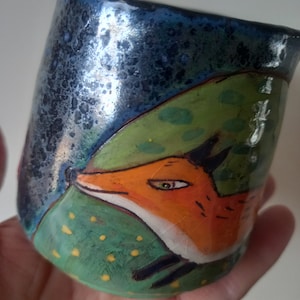 May include: A ceramic mug with a blue, green and orange design. The mug features a fox peeking out from behind a green leaf. The fox has a white face and orange fur.