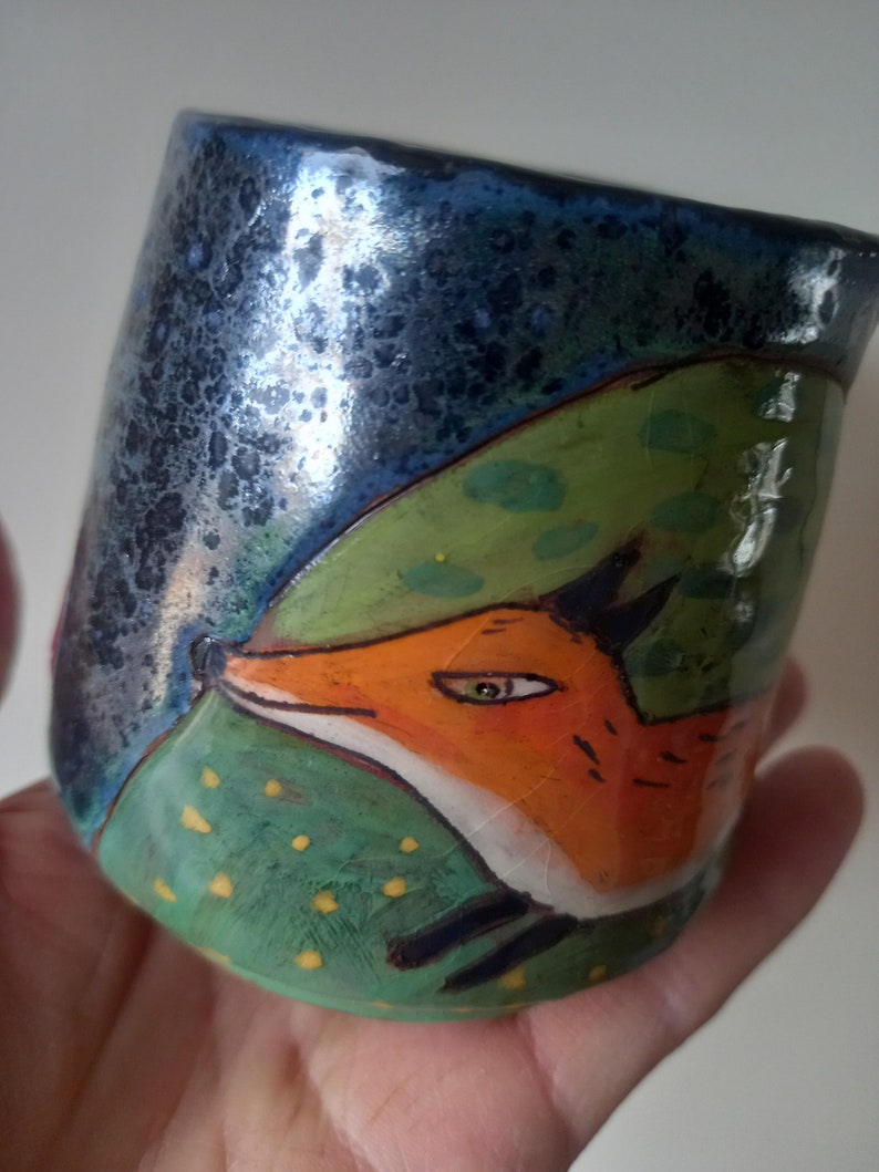 May include: A ceramic mug with a blue, green and orange design. The mug features a fox peeking out from behind a green leaf. The fox has a white face and orange fur.