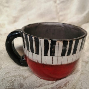 May include: A ceramic mug with a black handle and a red base. The mug is decorated with a black and white piano keyboard design.