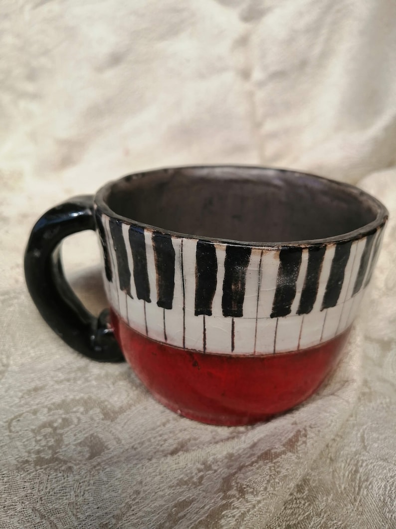 May include: A ceramic mug with a black handle and a red base. The mug is decorated with a black and white piano keyboard design.