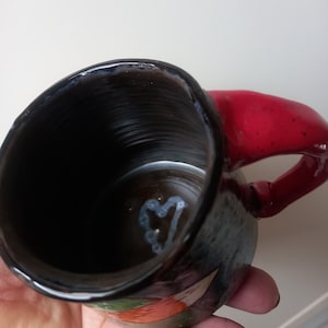 May include: A black ceramic mug with a red handle and a white heart design inside. The mug has a colourful floral pattern on the outside.
