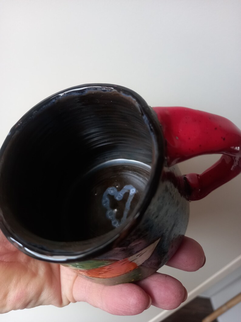 May include: A black ceramic mug with a red handle and a white heart design inside. The mug has a colourful floral pattern on the outside.
