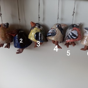 Puede incluir: Una colección de seis adornos de pájaros de cerámica, cada uno con patrones y diseños de colores únicos. Los pájaros están suspendidos por cuerdas, mostrando una variedad de colores que incluyen marrón, azul, amarillo y blanco. Cada pájaro tiene patas pequeñas y detalladas.