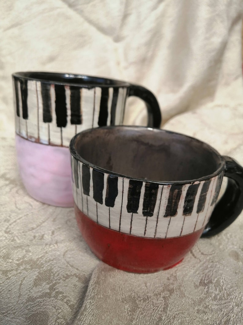 May include: Two ceramic mugs with black and white piano key designs. One mug has a pink base and the other has a red base.