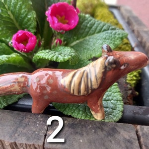 Puede incluir: Una figurita de caballo de cer&aacute;mica marr&oacute;n y beige con un acabado esmaltado. La figurita est&aacute; sentada sobre una superficie marr&oacute;n con plantas verdes en el fondo.
