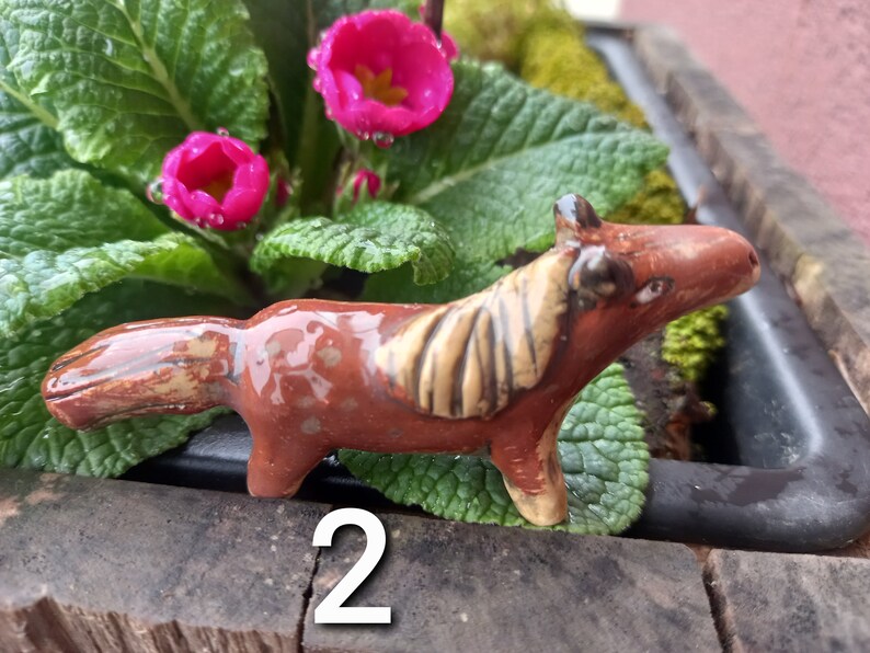 Puede incluir: Una figurita de caballo de cer&aacute;mica marr&oacute;n y beige con un acabado esmaltado. La figurita est&aacute; sentada sobre una superficie marr&oacute;n con plantas verdes en el fondo.