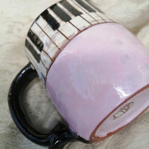May include: A ceramic mug with a black handle and a piano key design. The mug is painted with black and white piano keys on the top half and a light pink color on the bottom half.