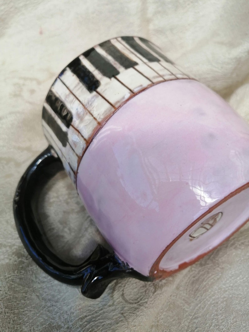 May include: A ceramic mug with a black handle and a piano key design. The mug is painted with black and white piano keys on the top half and a light pink color on the bottom half.