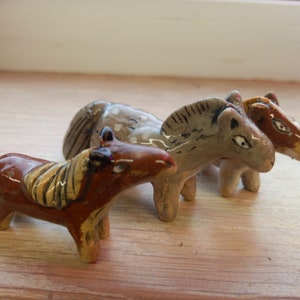 Puede incluir: Tres figuritas de caballos de cer&aacute;mica, una marr&oacute;n, una gris y una marr&oacute;n y blanca. Los caballos est&aacute;n de pie sobre una superficie de madera.