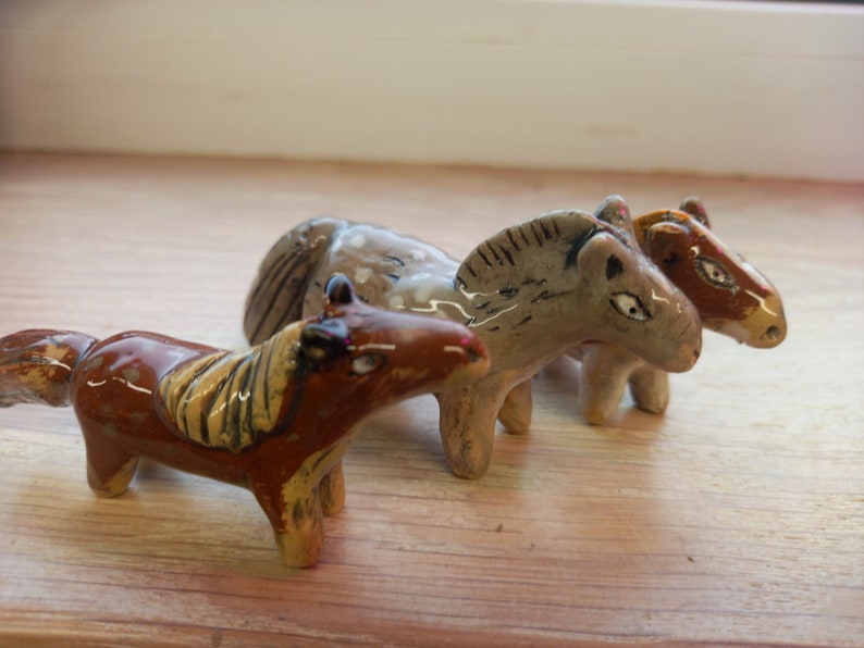 Puede incluir: Tres figuritas de caballos de cer&aacute;mica, una marr&oacute;n, una gris y una marr&oacute;n y blanca. Los caballos est&aacute;n de pie sobre una superficie de madera.