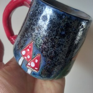 May include: A ceramic mug with a red handle and a dark blue glaze. The mug has a speckled finish and features a hand-painted design of red and white mushrooms.