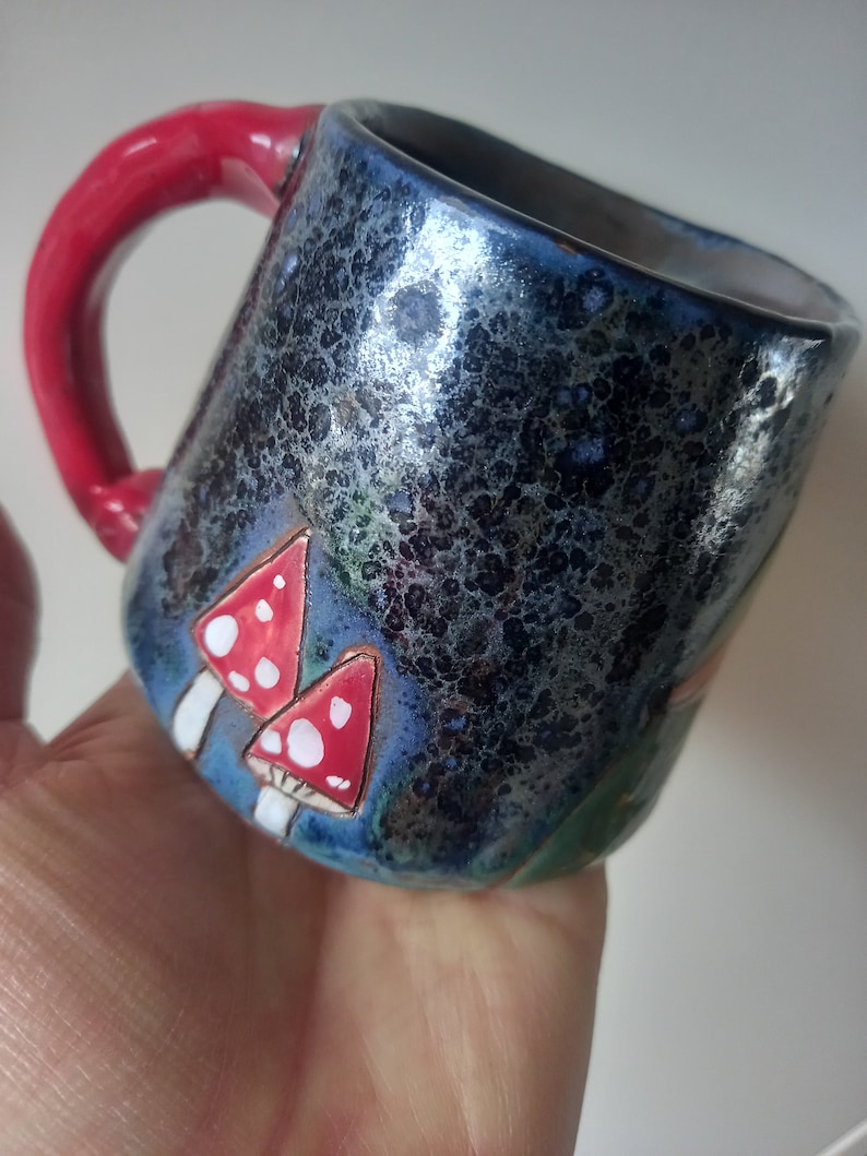 May include: A ceramic mug with a red handle and a dark blue glaze. The mug has a speckled finish and features a hand-painted design of red and white mushrooms.