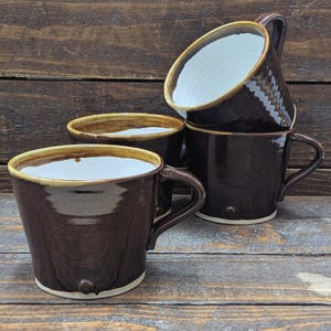 May include: Four brown ceramic mugs with a glossy finish are stacked. The mugs have a wide, slightly flared shape with a handle and a small decorative detail near the base. The rims are a lighter brown, and the interiors are white.