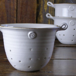 May include: White ceramic colander with two loop handles and small drainage holes. The colander has a slightly flared shape and a small logo on the side. The colander is stacked with two others, all with a rustic, handmade appearance.