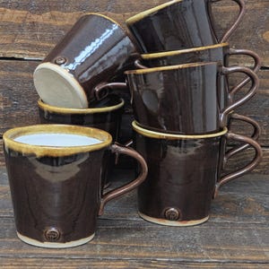 May include: A collection of six brown ceramic mugs with a glossy finish. Each mug features a rounded handle and a light tan rim. The mugs are stacked on a wooden surface.