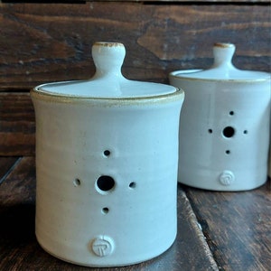 May include: Two off-white ceramic jars with lids. Each jar has a cylindrical shape, a small handle on the lid, and a series of small holes. The rims and handles have a gold-coloured trim. The jars are on a wooden surface.