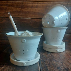 May include: White ceramic planters with drainage holes and matching saucers. One planter contains a wooden spoon and a pile of white material. The planters are on a wooden surface.