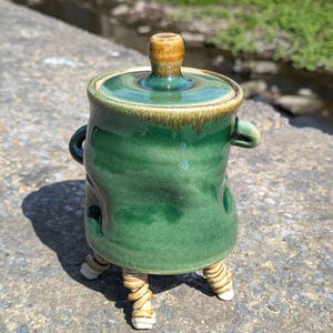 May include: A handcrafted ceramic container with a green glaze, a lid, and three twisted legs. The container has a small handle on each side and a brown knob on the lid. The container is approximately 10 cm tall.