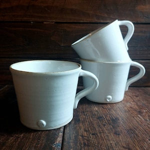 May include: Four white ceramic mugs with handles, three stacked and one in front. The mugs have a simple, rustic design with a slightly textured surface and a small decorative element near the base. The mugs are on a wooden surface.
