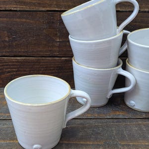 May include: Six white ceramic mugs with handles, stacked on a dark wooden surface. The mugs have a textured finish and a minimalist design. The rims are subtly accented with a light brown colour.