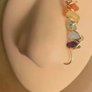 May include: A gold nose ring with a spiral design and seven colorful gemstones. The gemstones are red, orange, yellow, green, blue, purple, and clear.
