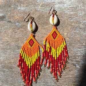 May include: A pair of long, beaded earrings with a shell charm at the top. The earrings are red, orange, and yellow.