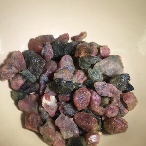 May include: A bowl filled with a variety of small, rough, green, pink, and black gemstones. The gemstones are tumbled and have a natural, unpolished look.