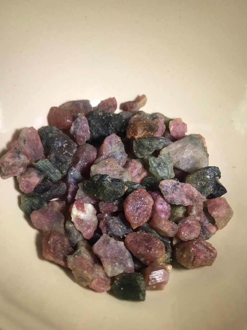 May include: A bowl filled with a variety of small, rough, green, pink, and black gemstones. The gemstones are tumbled and have a natural, unpolished look.