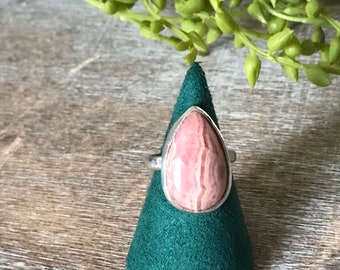 Puede incluir: Un anillo de plata con una piedra preciosa de color rosa y blanco en forma de lágrima. El anillo se muestra en un soporte de anillo verde.