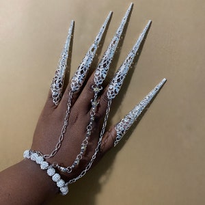 May include: A silver chain bracelet with a row of small, sparkly, round stones. The bracelet is connected to five silver finger rings with intricate designs. The rings are worn on the fingers of a hand, and each ring has a long, pointed, silver nail-like extension.