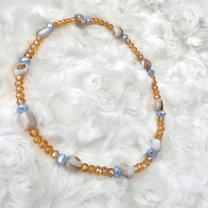 May include: A delicate necklace made with small, light brown glass beads and white and brown shell beads. The necklace also features small, light blue glass beads.