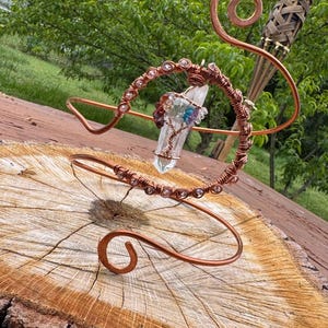 May include: A handcrafted copper wire sculpture featuring a clear quartz crystal with a wire-wrapped tree design. The sculpture has a spiral design and is adorned with small, colorful beads. A decorative torch is in the background.